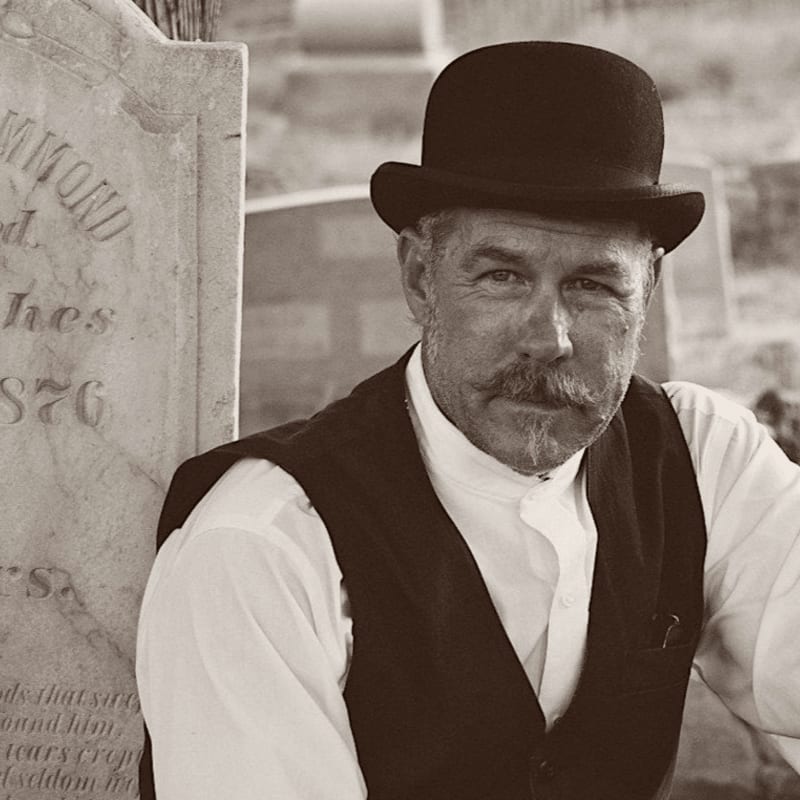 A man in a bowler hat and vest sits in front of a weathered tombstone, with a blurred, rustic background.