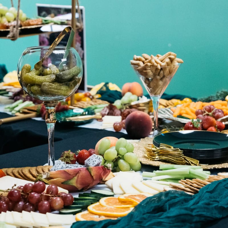 A table laden with a colorful assortment of fruits, cheeses, and crackers in the foreground is set against a teal wall in the background.