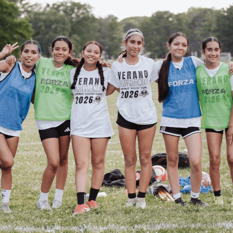 A group of young women in soccer uniforms stand on a grassy field with soccer equipment in the background.