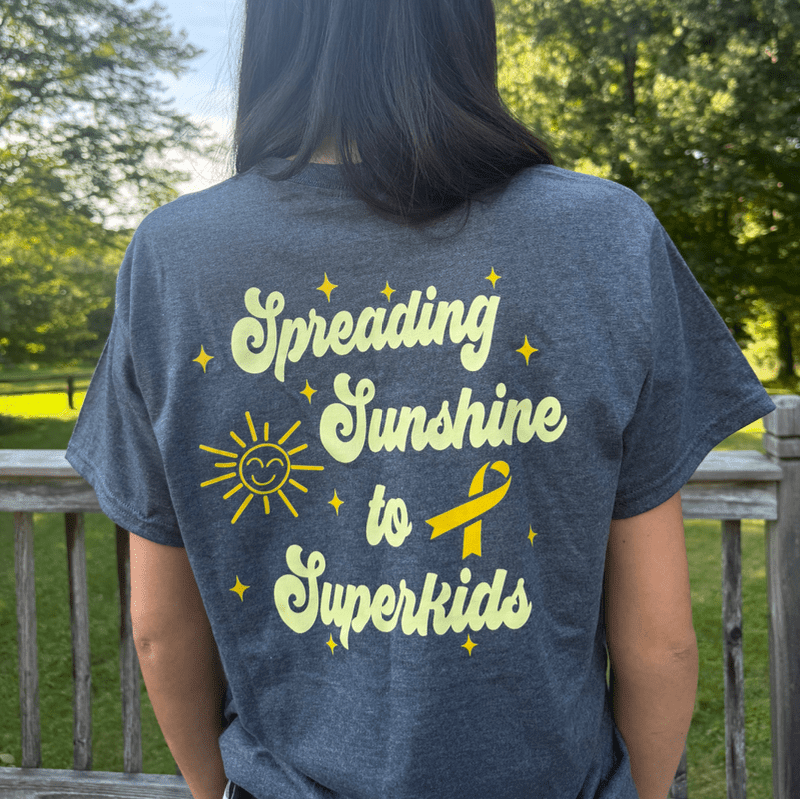 A person in a dark t-shirt with a yellow "Spreading Sunshine to Superkids" graphic stands in front of a wooden railing and blurred green trees.