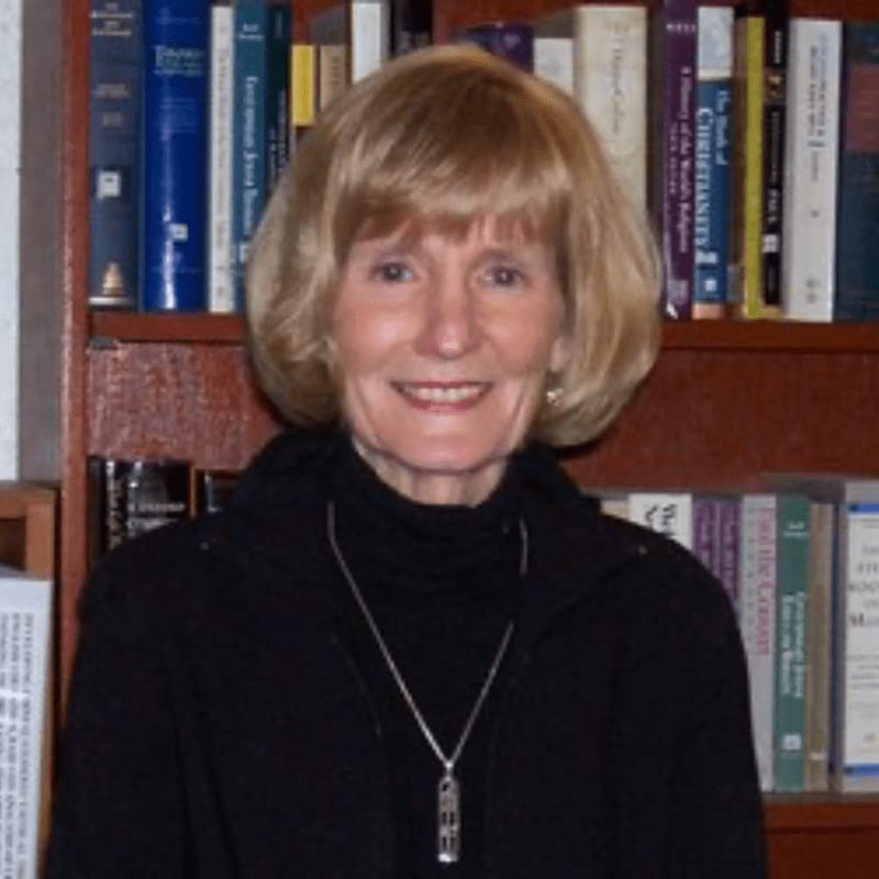 A smiling woman with blonde hair and a black turtleneck stands in front of a bookshelf filled with books.