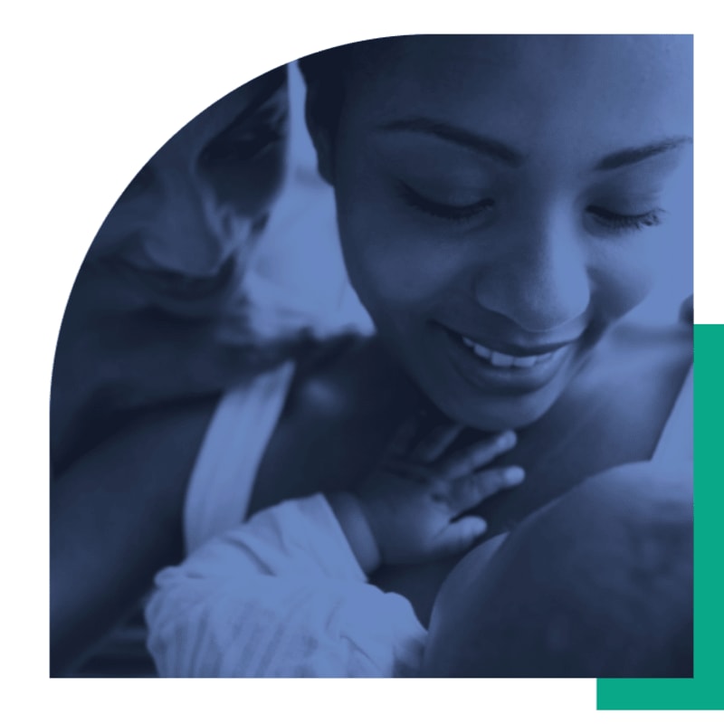 A mother smiles lovingly at her newborn baby in the foreground, with a blurred figure of another person in the background.