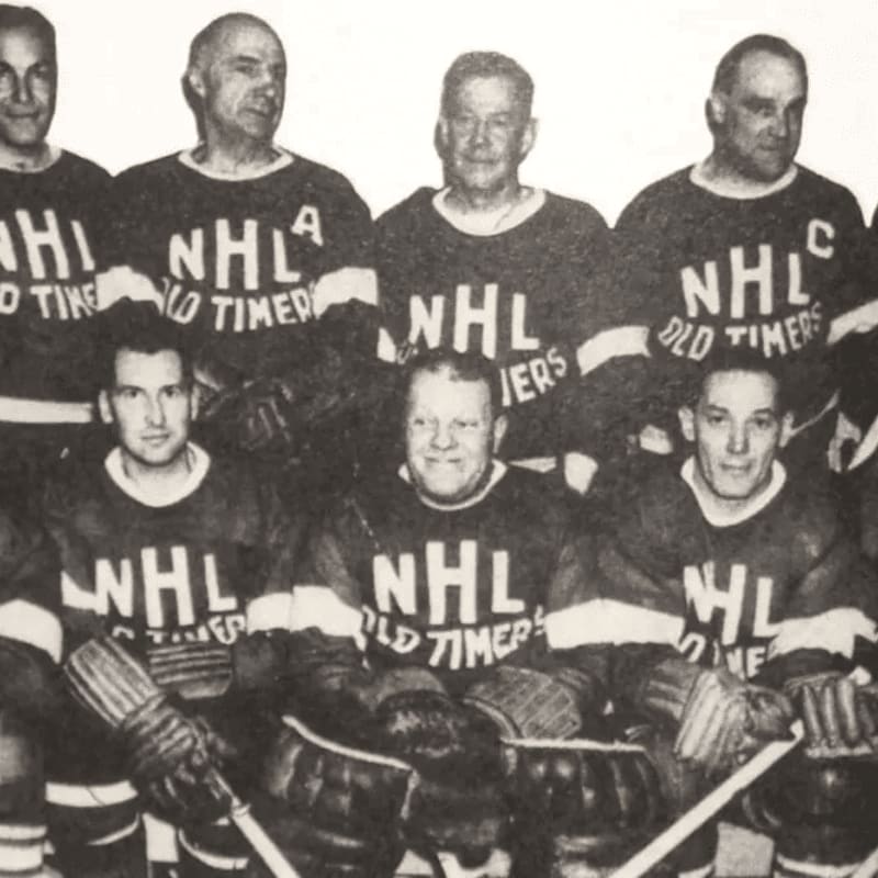 A group of men in NHL Old Timers jerseys pose for a photo, with some in the front row kneeling and others standing behind them.