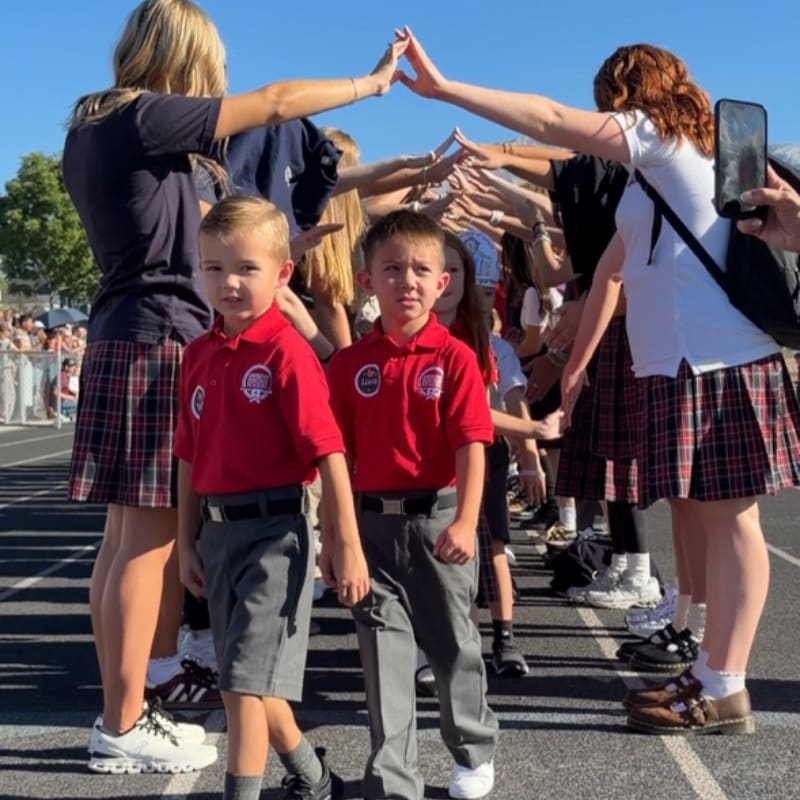 Two young boys in school uniforms walk through a tunnel of high-fiving students under a clear blue sky.
