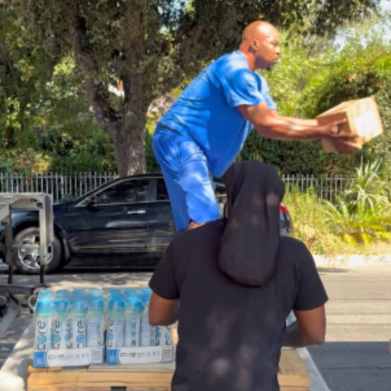 A man in blue scrubs stands on a vehicle handing a box to another person in a black shirt, with cases of bottled water in the foreground and trees and a fence in the background.