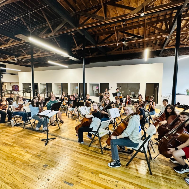 A group of young musicians with their instruments are seated in a large room with a wooden floor and exposed ceiling beams.