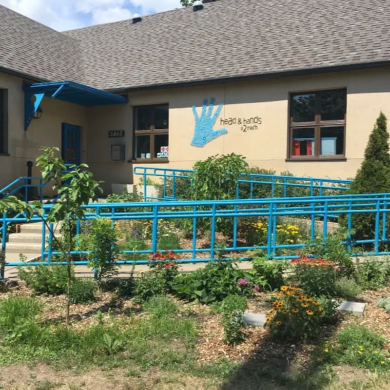 A vibrant garden with a blue ramp in the foreground leads to a building with a "head & hands" mural on its exterior.