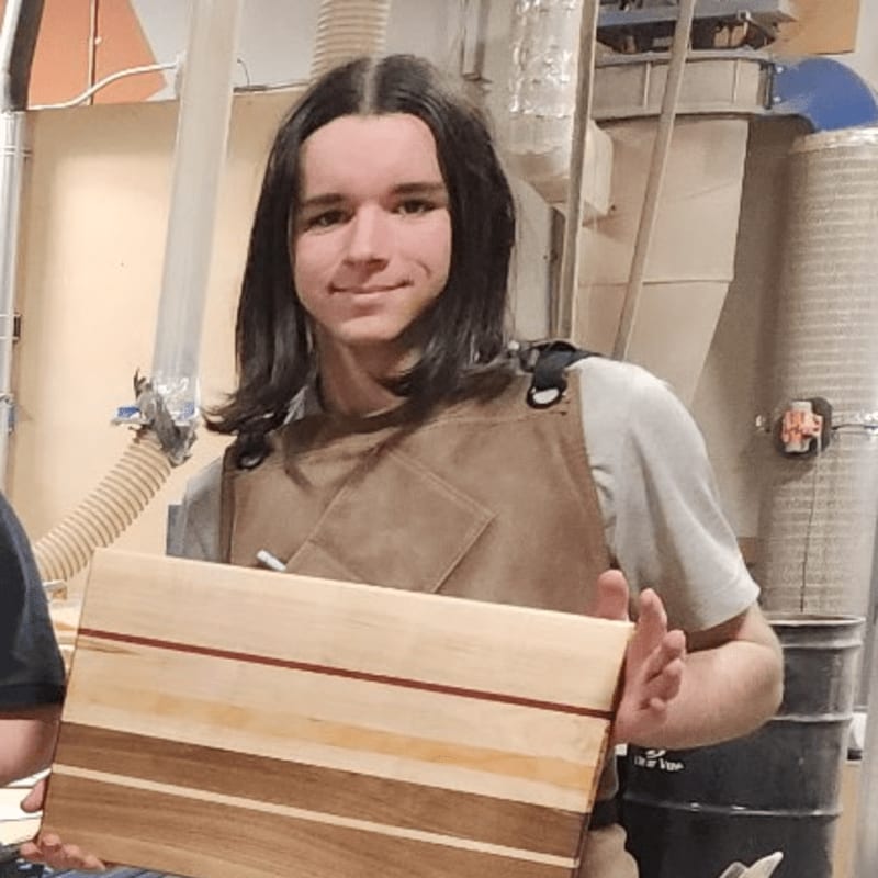 A young person in an apron proudly holds a handmade wooden cutting board in a workshop setting with dust collection equipment in the background.