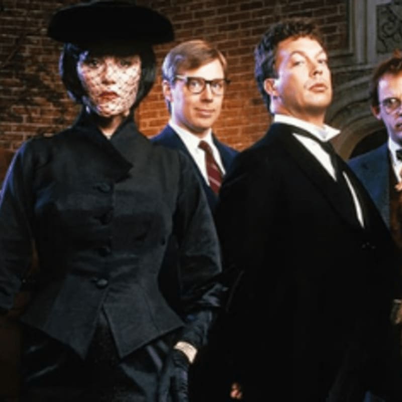 A woman in a black veil and hat stands in the foreground, with three men in formal attire behind her against a brick wall.
