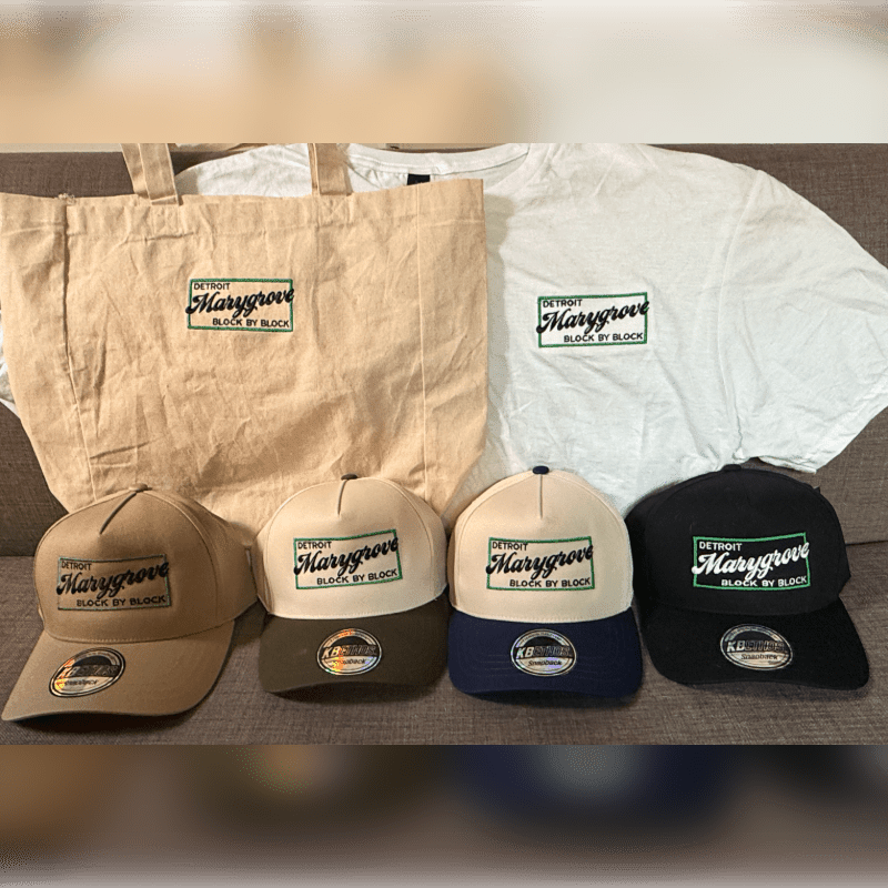 Four baseball caps are in the foreground, with a tote bag and t-shirt behind them, all featuring the "Detroit Marygrove Block by Block" logo.