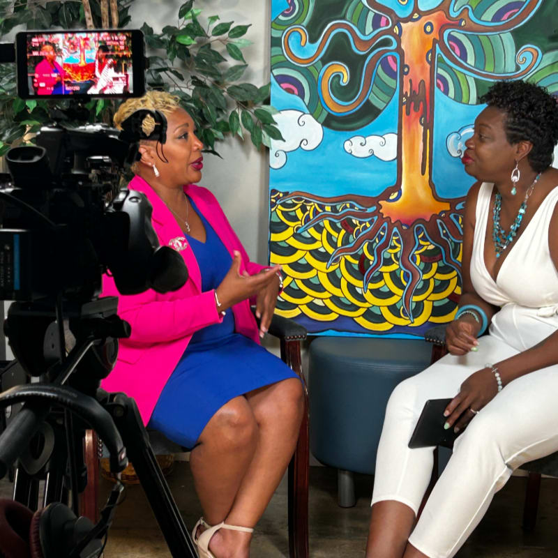 In the foreground, a camera is set up to film two women, one in a pink jacket and blue dress, the other in a white outfit, seated in front of a colorful tree painting.