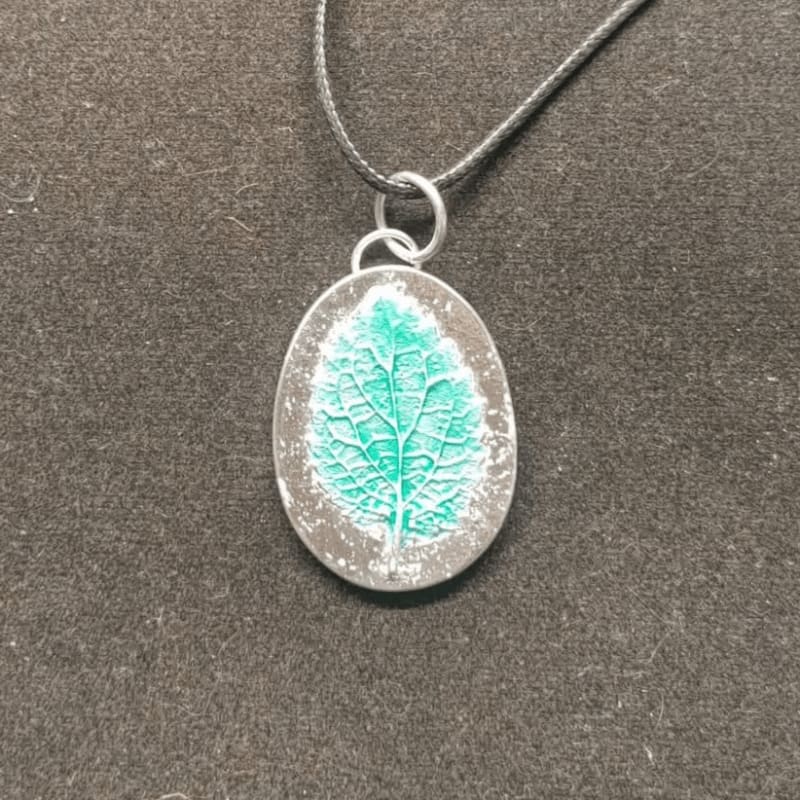 A teal leaf pendant with a textured surface hangs from a black cord against a dark, textured background.