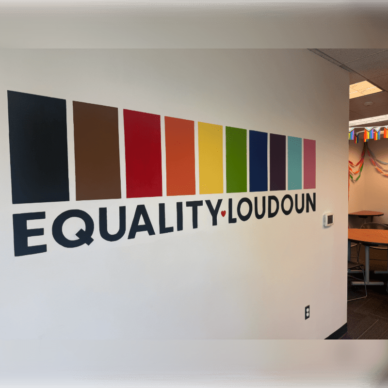 A colorful pride flag-inspired logo with the words "EQUALITY LOUDOUN" is prominently displayed on a white wall in the foreground, while the background reveals a glimpse of a room with tables and rainbow bunting.