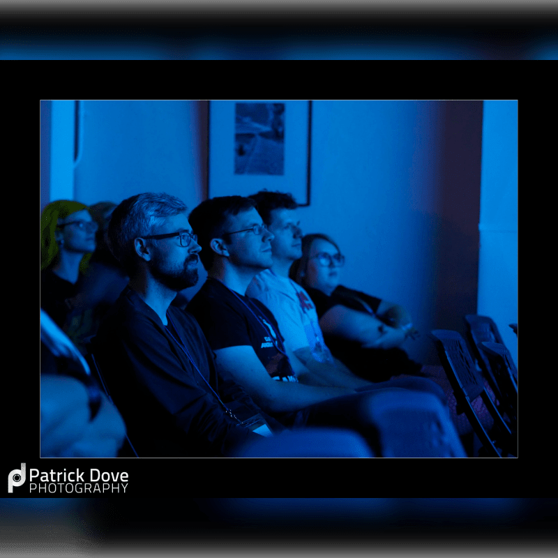 A group of people with focused expressions watch something in a dimly lit, blue-toned room.