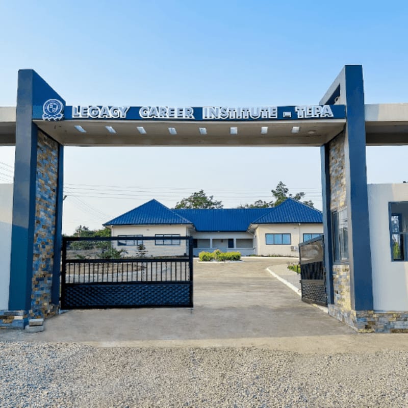 A wide, open gate with a gravel driveway leads to a modern building with blue roofs, all under a clear blue sky.