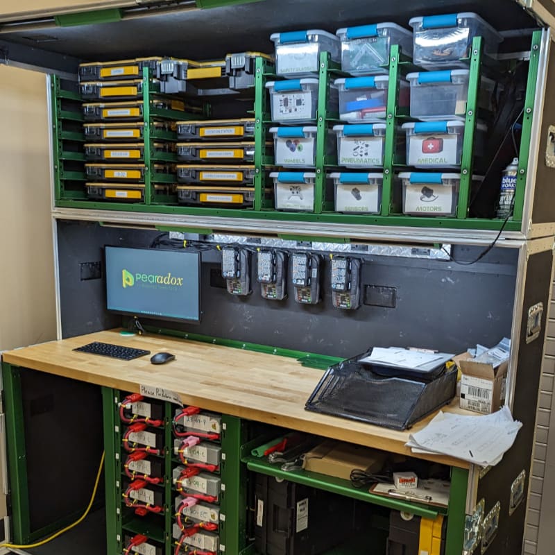 A workbench with a computer and storage bins is set up inside a large, open case with shelves of equipment above and below.