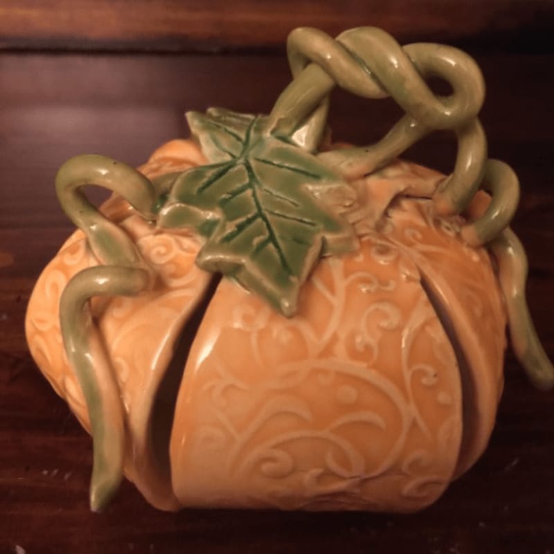 A decorative ceramic pumpkin with swirling vine details sits on a dark wooden surface, with a blurred wooden background.