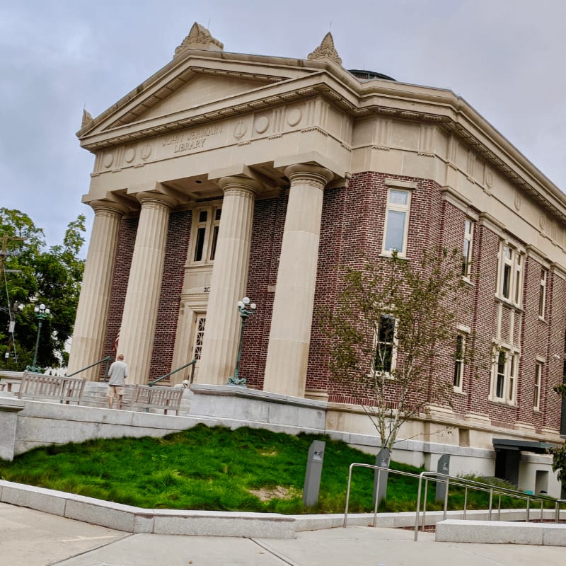 A person walks up the steps of a grand brick library with large columns, set against a cloudy sky.