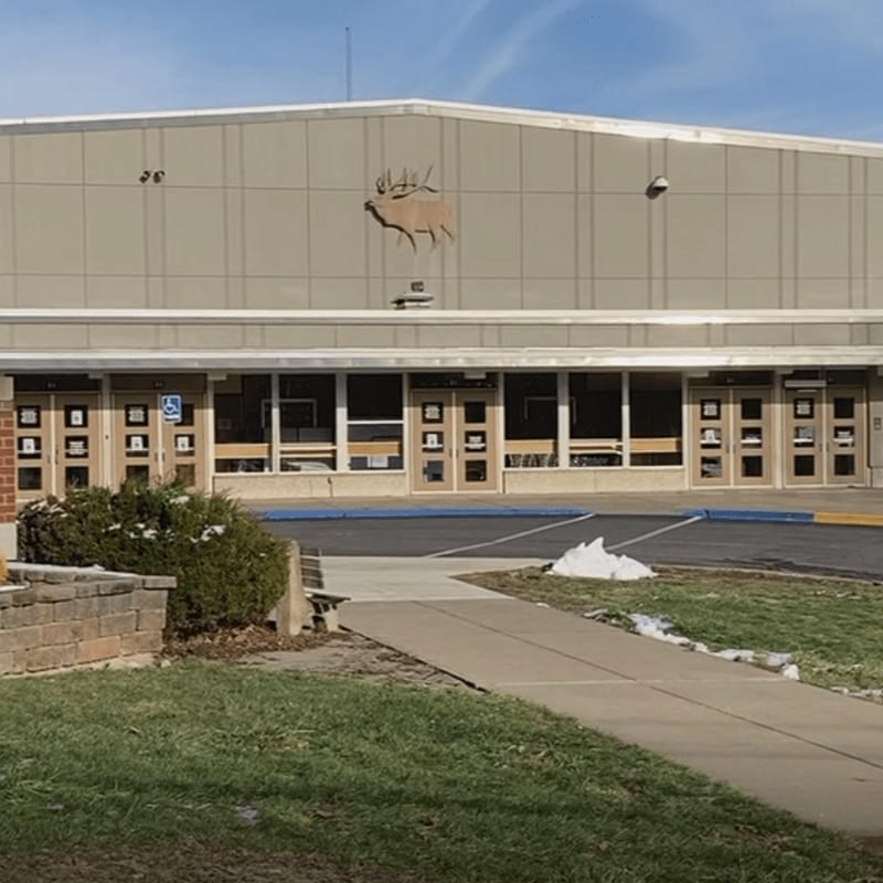 A concrete building with an elk emblem is visible behind a walkway and grassy area with patches of snow.