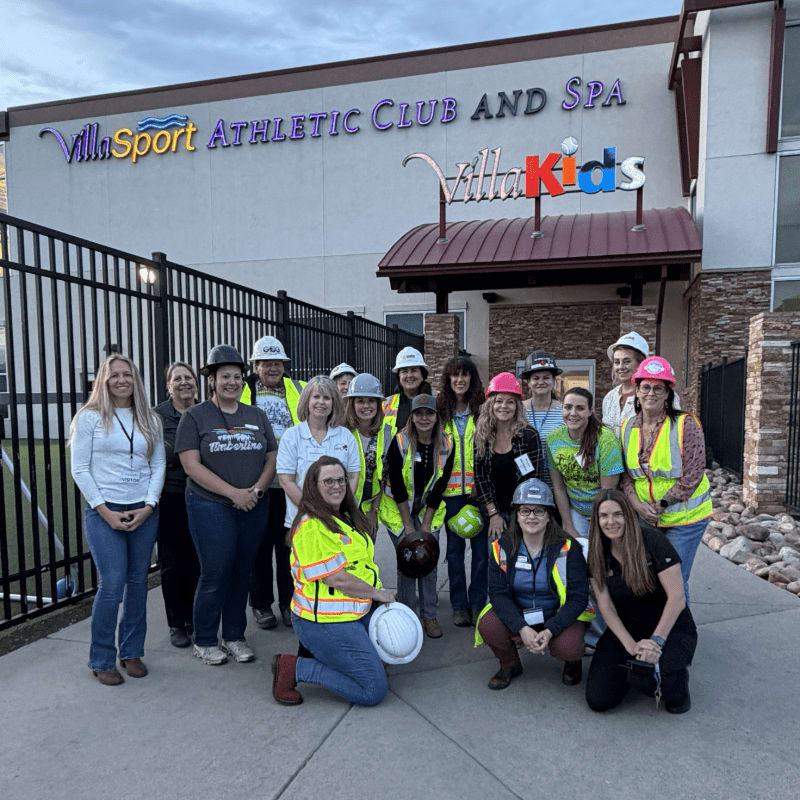 A group of women in hard hats and safety vests pose in front of the Villa Sport Athletic Club and Spa, with a black metal fence in the foreground.