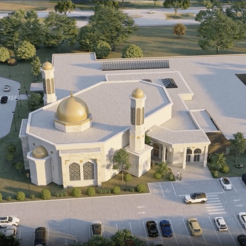 A modern mosque with a golden dome and minarets stands in a parking lot surrounded by trees and a road in the background.