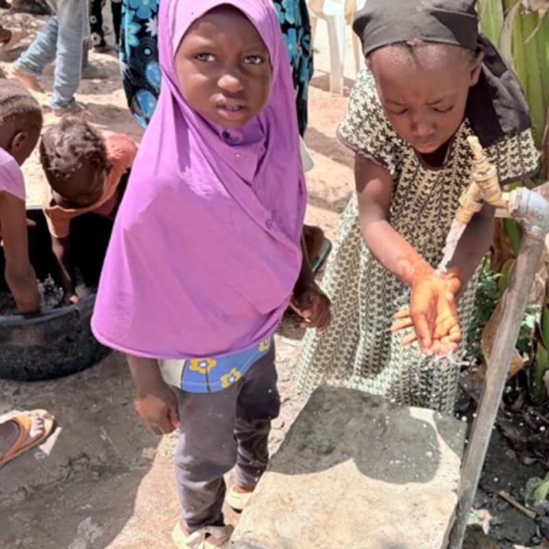 Two young children, one in a purple hijab and the other washing their hands at a faucet, stand in a dusty outdoor area with other children and a building in the background.