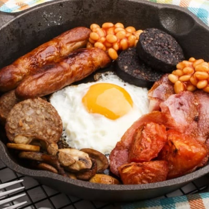 A hearty full English breakfast with sausages, bacon, eggs, beans, black pudding, and mushrooms fills a cast-iron skillet against a checkered cloth background.