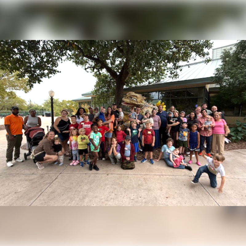 A large group of people, including many children, pose for a photo in front of a building with a large tree overhead.