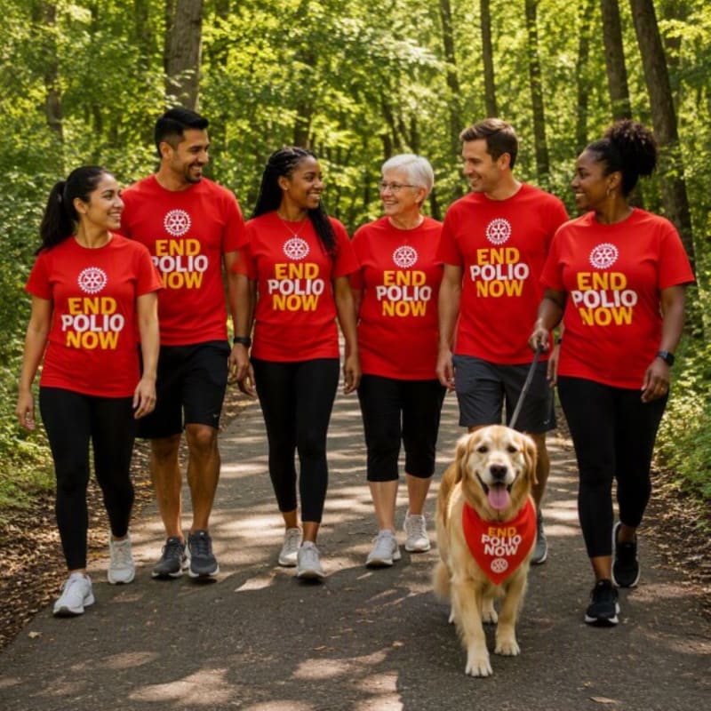 A group of six people wearing "End Polio Now" t-shirts and a golden retriever wearing a matching bandana walk on a wooded path.