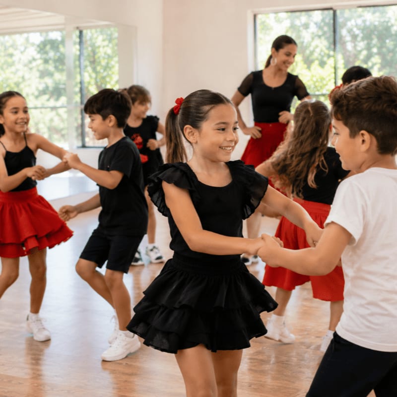 Children in black and red dance attire twirl in a bright studio with a teacher observing in the background.
