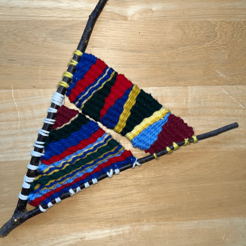 A colorful woven craft made with yarn on a triangular frame of sticks rests on a wooden surface.