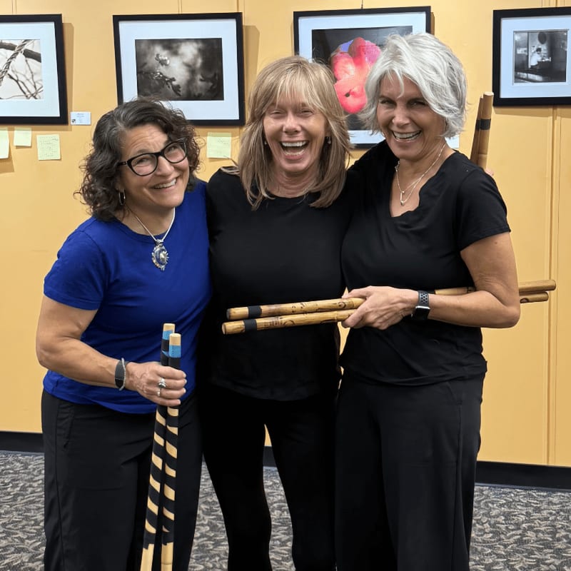 Three smiling women in black tops and pants hold wooden staffs in front of framed artwork on a yellow wall.