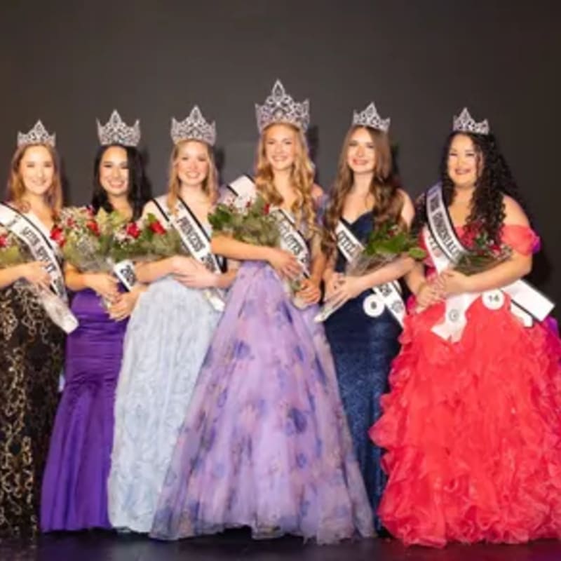 Six young women in formal gowns and crowns stand holding bouquets against a dark background.