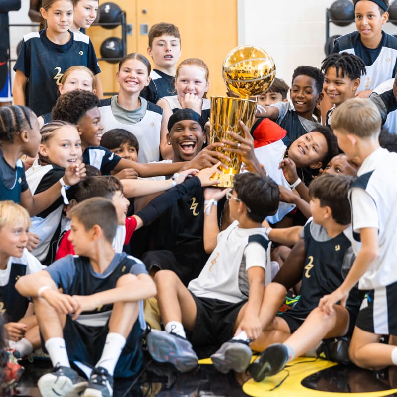 A smiling man in a black jersey holds a championship trophy surrounded by excited children in a gym.