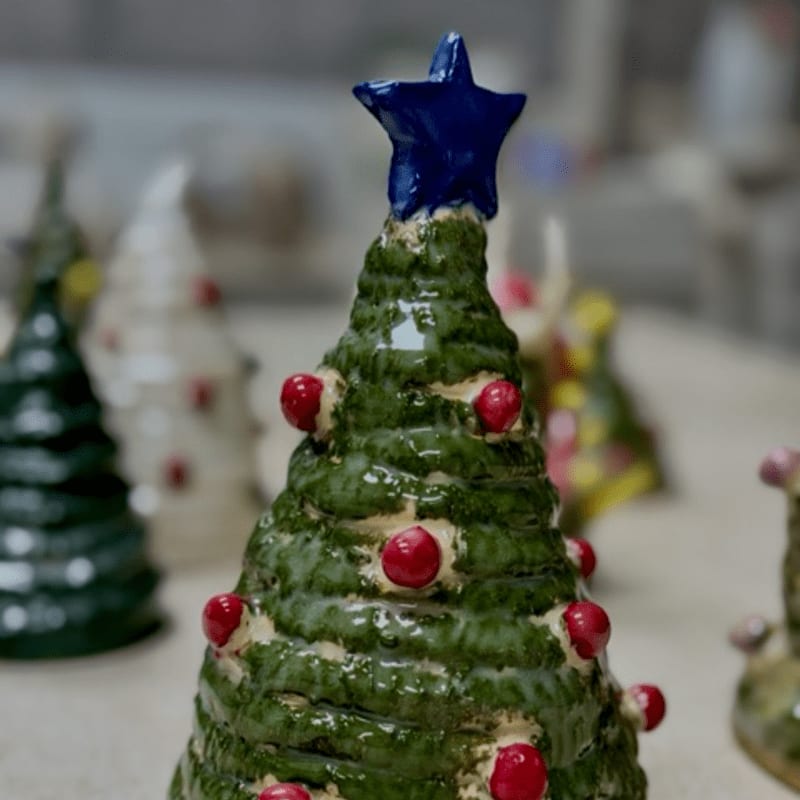 A green ceramic Christmas tree with red ornaments and a blue star sits in the foreground, with other colorful ceramic trees blurred in the background.