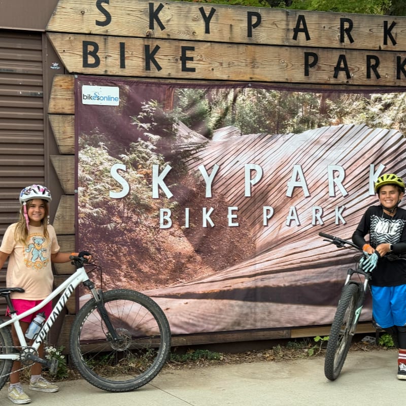 Two young girls with their bikes stand in front of a large sign for Skypark Bike Park, which is set against a backdrop of trees and a dirt trail.