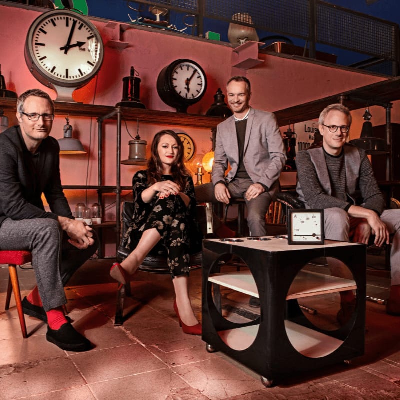 Four people are seated in a room filled with clocks, with a modern table in the foreground.