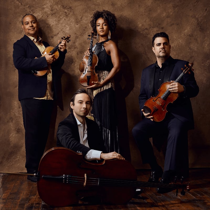 Four musicians, three holding violins and one with a cello, pose against a textured brown background.