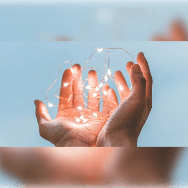Cupped hands hold a string of glowing fairy lights against a soft blue sky.