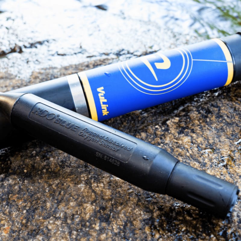 Two black and blue sensors rest on a textured rock surface with a blurred watery background.