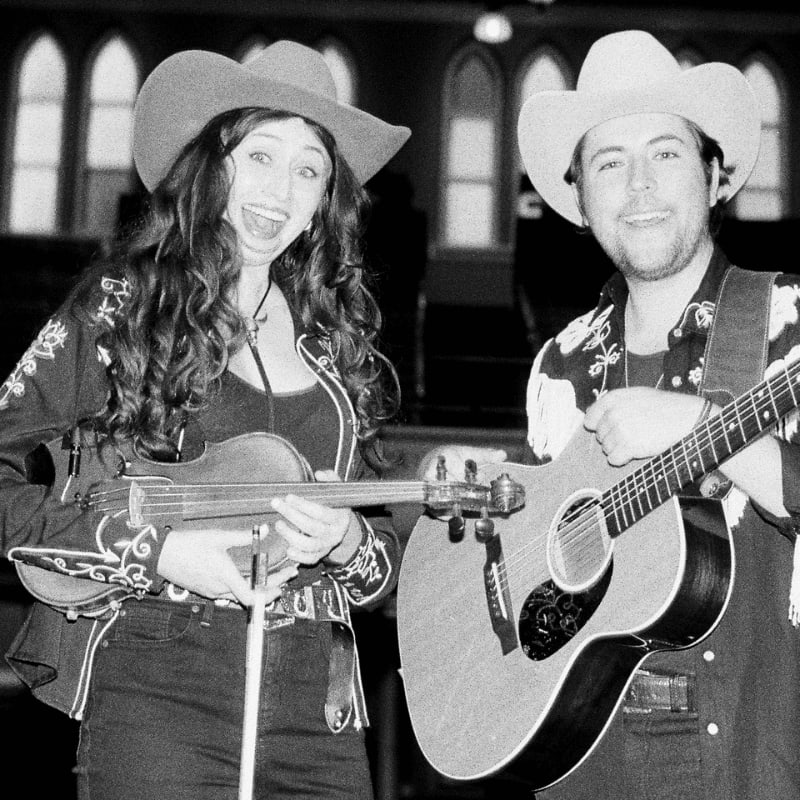 A woman in a cowboy hat and embroidered jacket plays the violin while a man in a cowboy hat and patterned shirt strums a guitar, with arched windows visible in the dimly lit background.