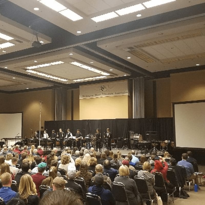 A large audience fills the foreground of a convention hall, facing a stage where musicians perform in front of a banner for the "Washington State Thespian Association" in the background.