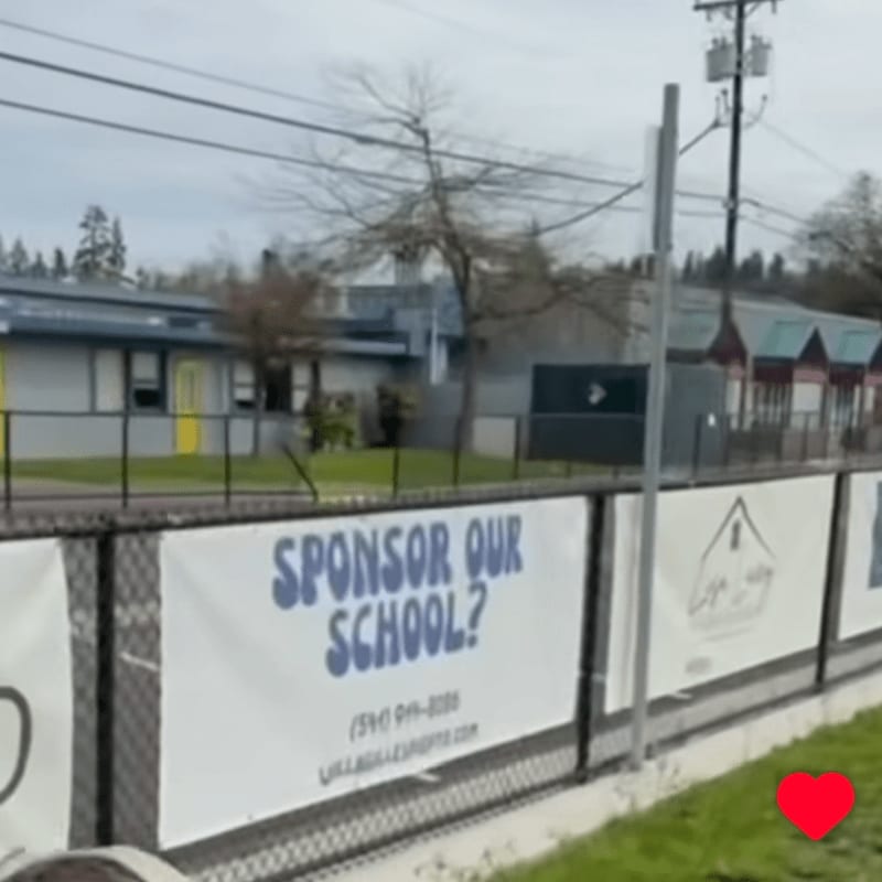 A fence with a "Sponsor Our School?" banner in the foreground separates a grassy area from a school building with a playground in the background.