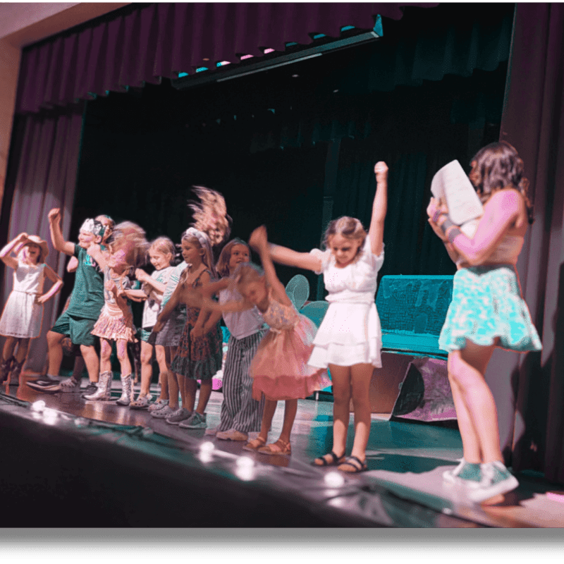A group of children in colorful outfits are celebrating on a stage with a dark curtain and spotlights in the background.