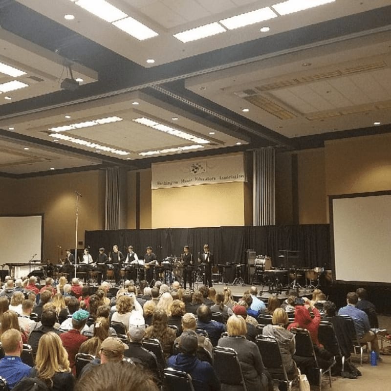 A large audience fills the foreground of a convention hall, facing a stage where musicians perform in front of a banner for the "Washington State Thespian Association" in the background.
