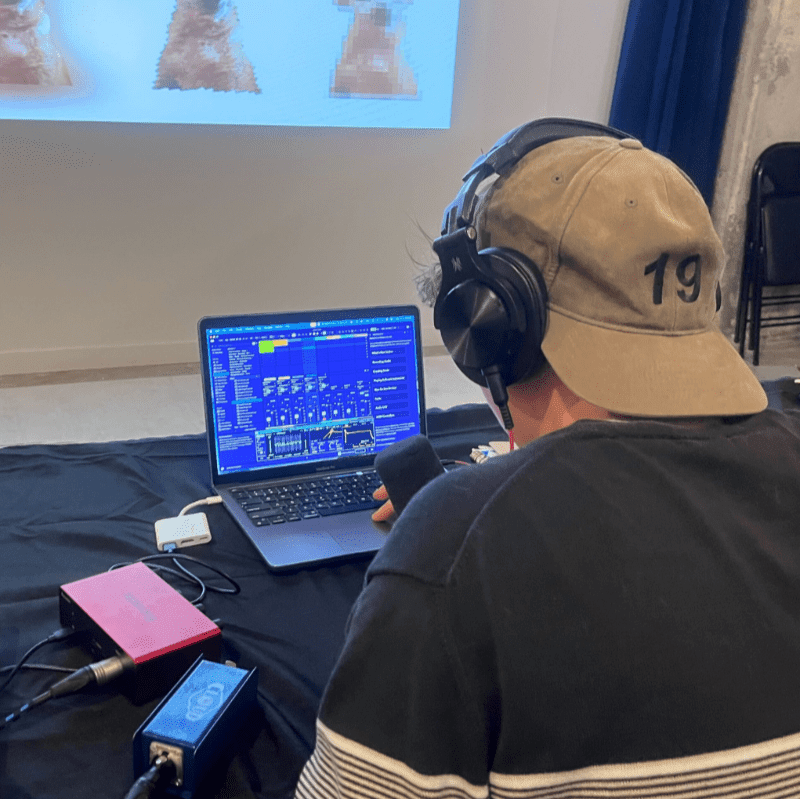 A person wearing headphones and a baseball cap is working on a laptop with audio equipment in the foreground, while a projected image is visible on a screen in the background.