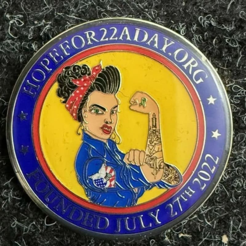 A coin with a Rosie the Riveter-style woman flexing her bicep, featuring the text "HOPEFOR22ADAY.ORG FOUNDED JULY 27TH 2022" around the edge, rests on a dark, textured surface.