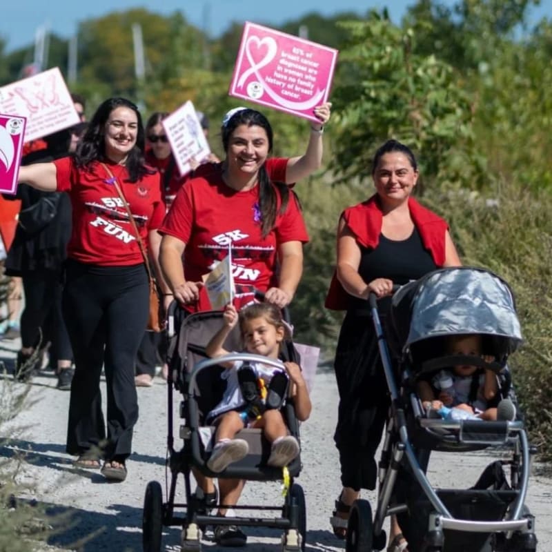 Women in red shirts push strollers with children along a path, while others in the background hold pink signs.