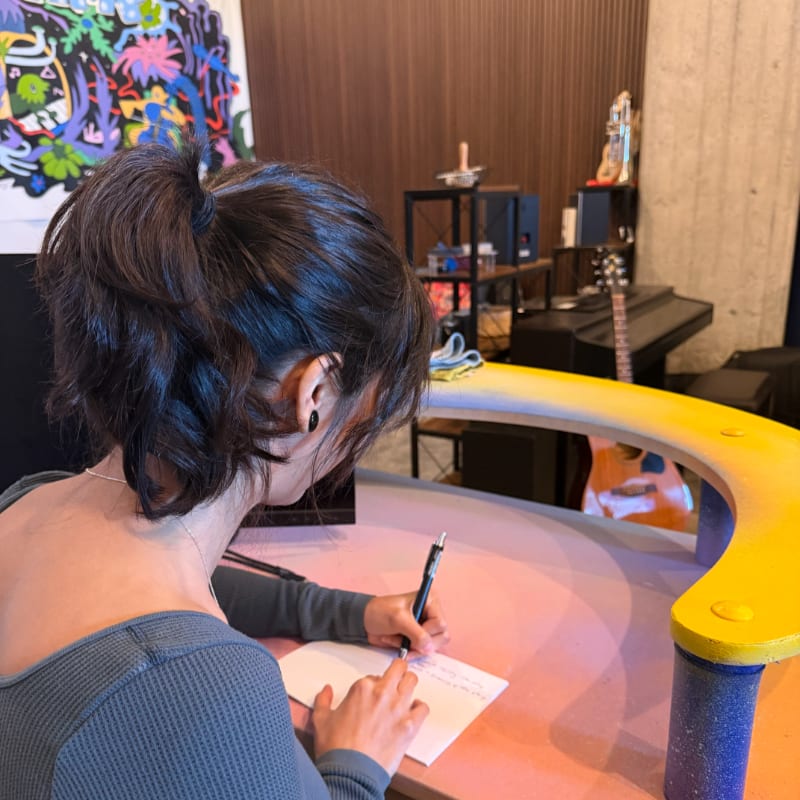 A woman with her back to the camera writes on a piece of paper at a colorful desk, with a piano and abstract art visible in the background.