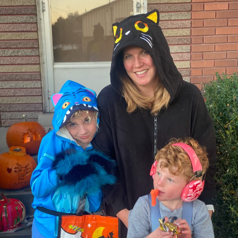 A woman in a black cat costume smiles with two children in costumes, one a blue monster and the other wearing headphones, in front of a house with carved pumpkins.